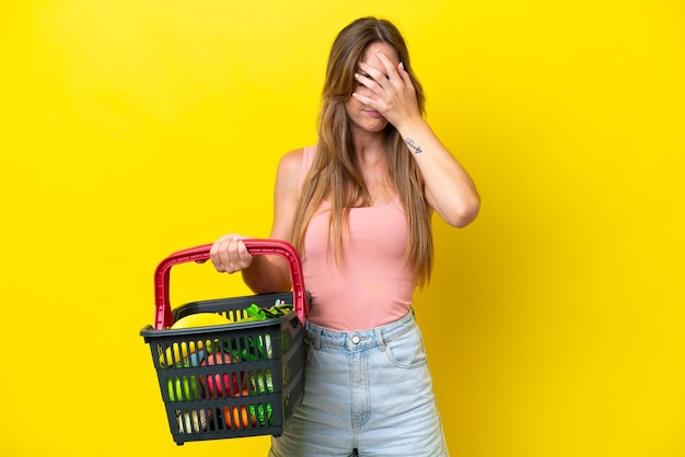 Jovem mulher caucasiana segurando uma cesta de compras cheia de comida isolada em fundo amarelo com expressão cansada e doente