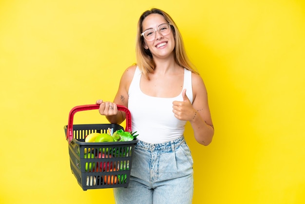 Jovem mulher caucasiana segurando uma cesta de compras cheia de comida isolada em fundo amarelo dando um polegar para cima gesto