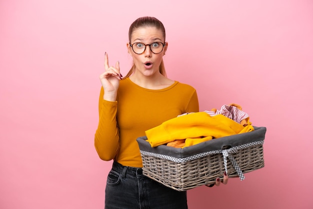 Jovem mulher caucasiana segurando uma cesta de roupas isolada em fundo rosa com a intenção de perceber a solução enquanto levanta um dedo