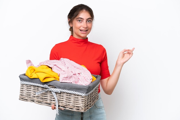 Jovem mulher caucasiana segurando uma cesta de roupas isolada no fundo branco, apontando o dedo para o lado