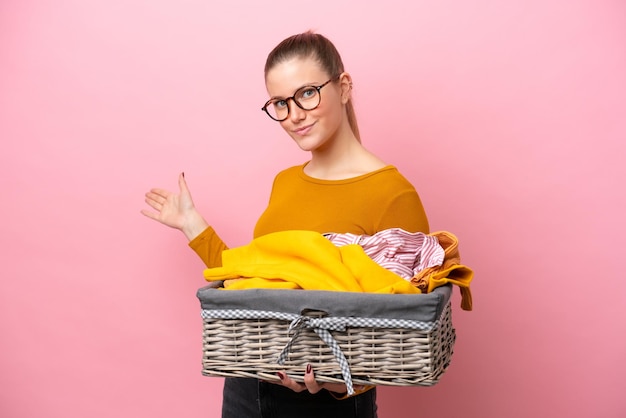 Jovem mulher caucasiana segurando uma cesta de roupas isolada no fundo rosa, estendendo as mãos para o lado para convidar para vir