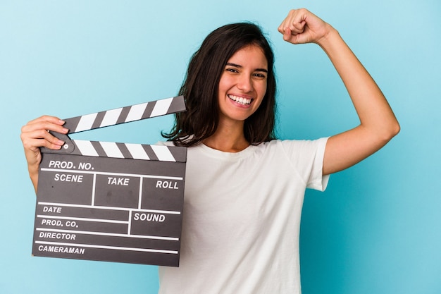 Jovem mulher caucasiana segurando uma claquete isolada no fundo azul, levantando o punho após uma vitória, o conceito de vencedor.