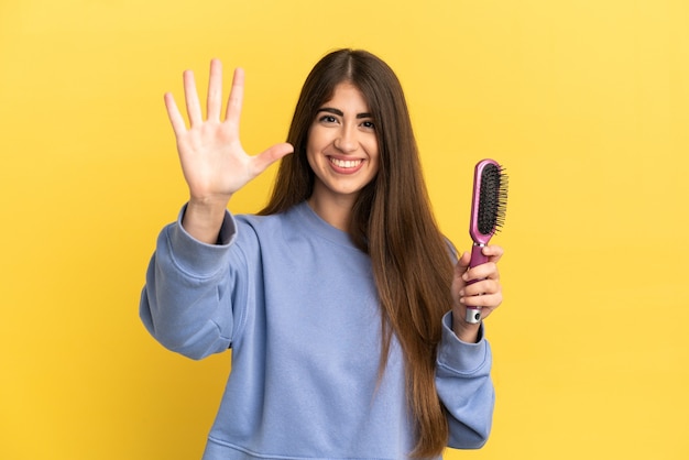 Jovem mulher caucasiana segurando uma escova de cabelo isolada em um fundo azul, contando cinco com os dedos