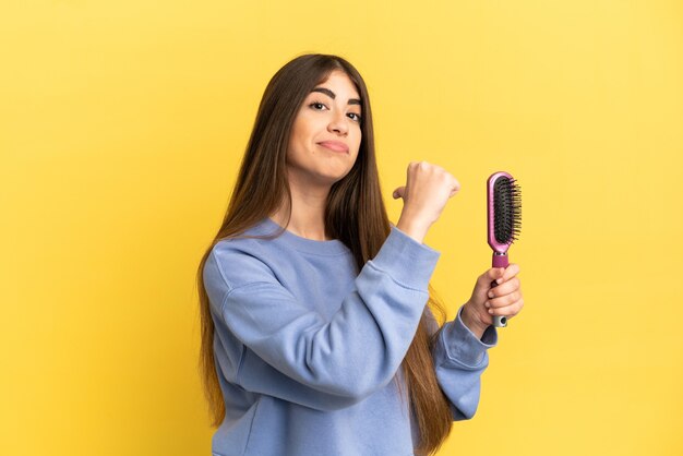 Jovem mulher caucasiana segurando uma escova de cabelo isolada em um fundo azul, orgulhosa e satisfeita