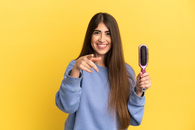 Jovem mulher caucasiana segurando uma escova de cabelo isolada em um fundo azul surpresa e apontando para a frente