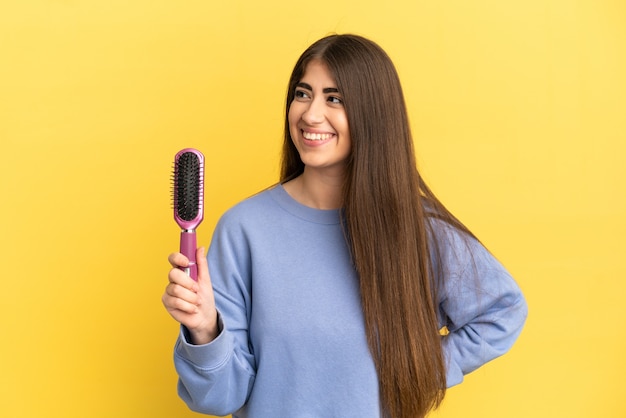 Jovem mulher caucasiana segurando uma escova de cabelo isolada na parede azul, olhando para o lado