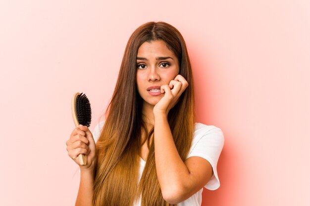 Jovem mulher caucasiana, segurando uma escova de cabelo, roendo as unhas, nervosa e muito ansiosa.