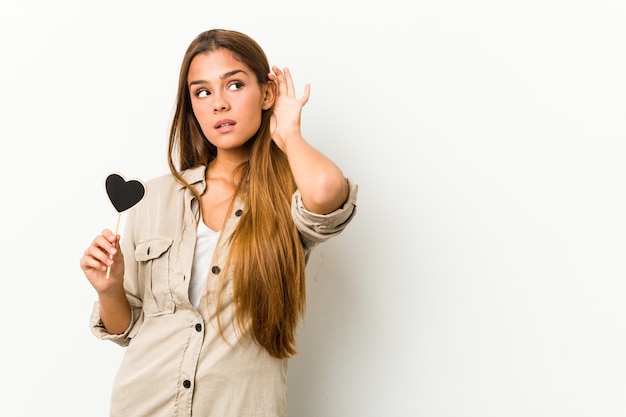 Jovem mulher caucasiana, segurando uma forma de coração, tentando ouvir uma fofoca.