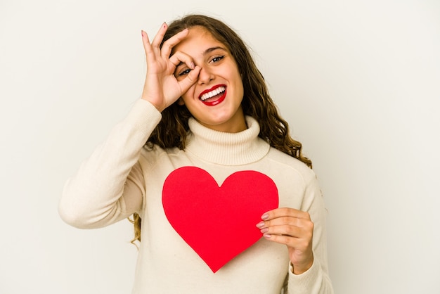 Jovem mulher caucasiana segurando uma forma de dia dos namorados coração isolada animado mantendo o gesto ok no olho.