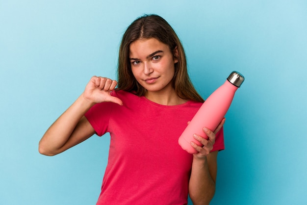 Jovem mulher caucasiana segurando uma garrafa de água isolada em um fundo azul, mostrando um gesto de antipatia, polegares para baixo. Conceito de desacordo.