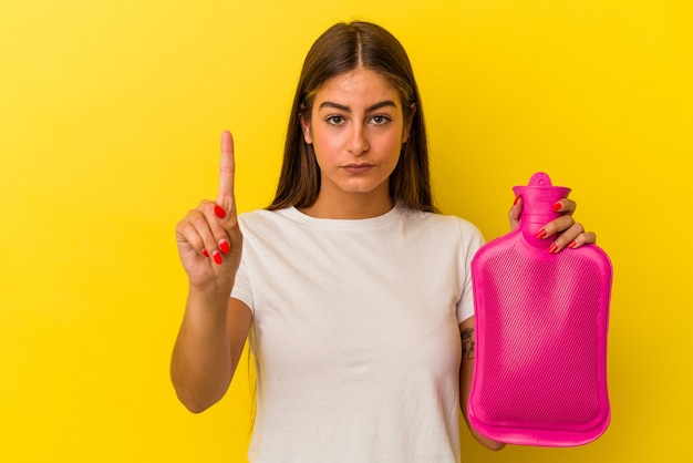 Jovem mulher caucasiana, segurando uma garrafa de água quente isolada no fundo amarelo, mostrando o número um com o dedo.