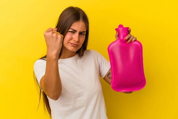 Jovem mulher caucasiana, segurando uma garrafa de água quente isolada no fundo amarelo, mostrando o punho para a câmera, expressão facial agressiva.