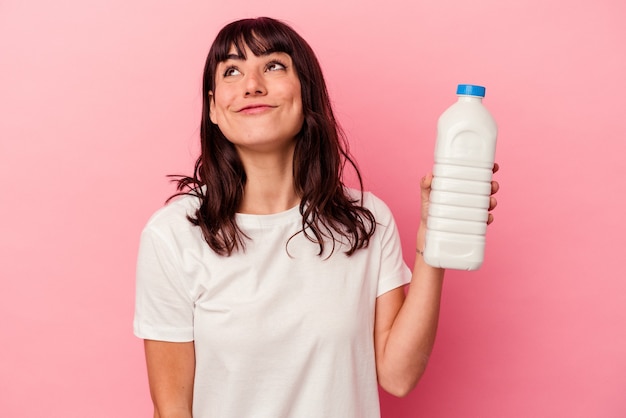 Jovem mulher caucasiana segurando uma garrafa de leite isolada na parede rosa, sonhando em alcançar objetivos e propósitos