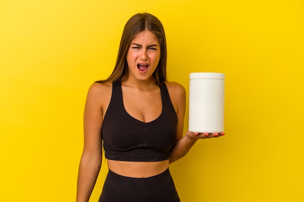 Jovem mulher caucasiana segurando uma garrafa de proteína isolada em fundo amarelo, gritando com muita raiva e agressividade.