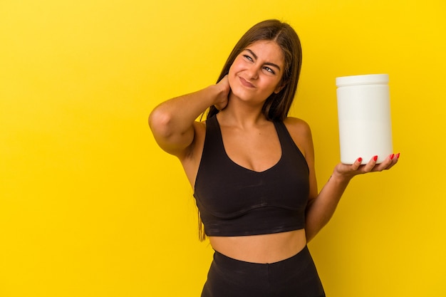 Jovem mulher caucasiana, segurando uma garrafa de proteína isolada em um fundo amarelo, tocando a parte de trás da cabeça, pensando e fazendo uma escolha.