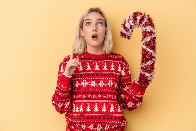 Jovem mulher caucasiana, segurando uma grande vara de Natal isolada em um fundo amarelo, apontando para cima com a boca aberta.