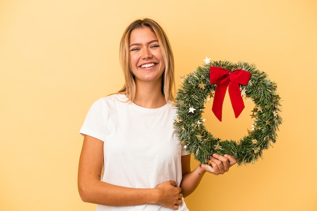 Jovem mulher caucasiana, segurando uma guirlanda de Natal isolada em fundo amarelo, rindo e se divertindo.