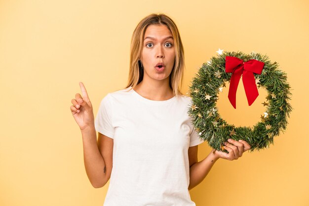 Jovem mulher caucasiana segurando uma guirlanda de Natal isolada em um fundo amarelo apontando para o lado