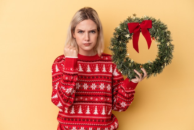 Jovem mulher caucasiana segurando uma guirlanda de Natal isolada em um fundo amarelo, mostrando o punho para a câmera, expressão facial agressiva.