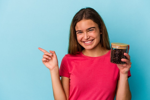 Jovem mulher caucasiana segurando uma jarra de café isolada em um fundo azul, sorrindo e apontando para o lado, mostrando algo no espaço em branco.