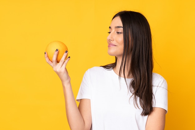 Jovem mulher caucasiana, segurando uma laranja sobre parede amarela