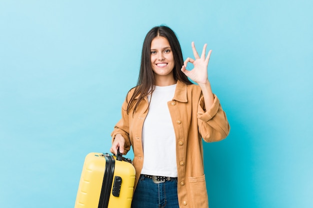 Jovem mulher caucasiana, segurando uma mala alegre e confiante, mostrando um gesto de ok.