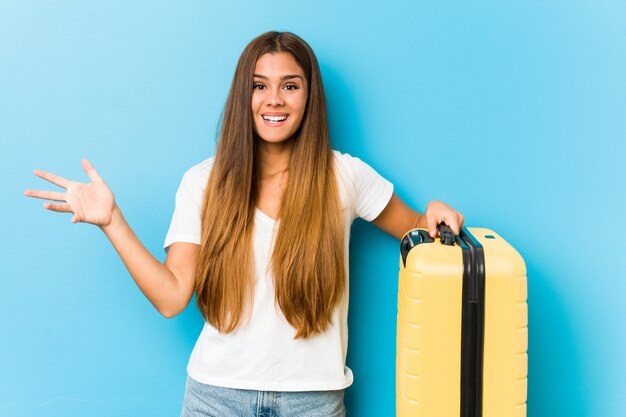 Jovem mulher caucasiana, segurando uma mala de viagem, recebendo uma agradável surpresa, animada e levantando as mãos.