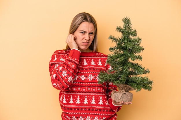 Jovem mulher caucasiana segurando uma pequena árvore de natal isolada em fundo amarelo, cobrindo os ouvidos com as mãos.