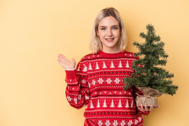 Jovem mulher caucasiana segurando uma pequena árvore de Natal isolada em fundo amarelo, mostrando um espaço de cópia na palma da mão e segurando a outra mão na cintura.