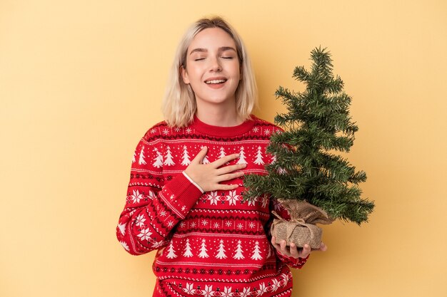 Jovem mulher caucasiana segurando uma pequena árvore de Natal isolada em fundo amarelo ri alto, mantendo a mão no peito.