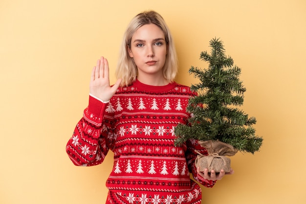 Jovem mulher caucasiana segurando uma pequena árvore de Natal isolada em um fundo amarelo em pé com a mão estendida, mostrando a placa de pare, impedindo você.