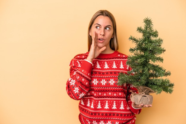 Jovem mulher caucasiana segurando uma pequena árvore de Natal isolada em um fundo amarelo está contando uma notícia secreta de travagem e olhando para o lado