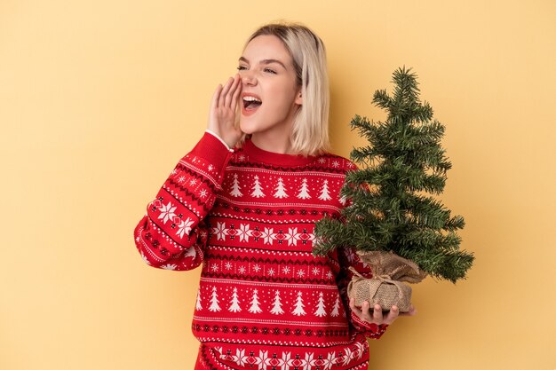 Jovem mulher caucasiana, segurando uma pequena árvore de Natal isolada em um fundo amarelo, gritando e segurando a palma da mão perto da boca aberta.