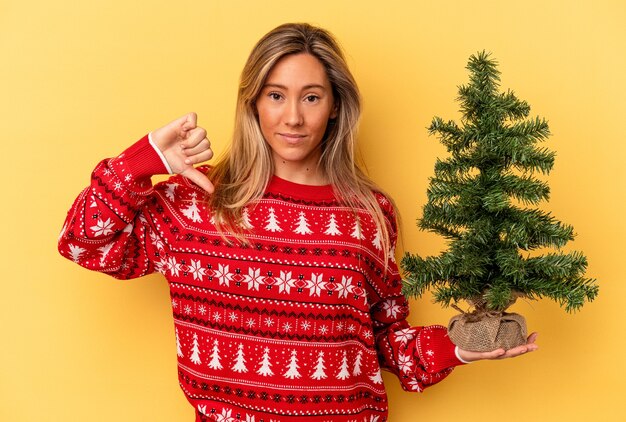 Jovem mulher caucasiana segurando uma pequena árvore de Natal isolada em um fundo amarelo, mostrando um gesto de antipatia, polegares para baixo. Conceito de desacordo.