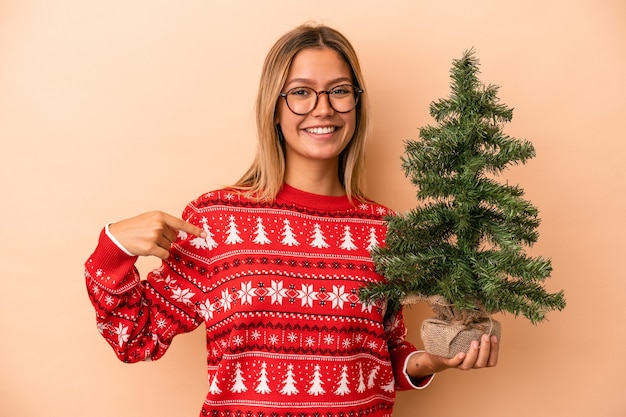 Jovem mulher caucasiana segurando uma pequena árvore de Natal isolada em um fundo bege pessoa apontando com a mão para um espaço de cópia de camisa, orgulhosa e confiante
