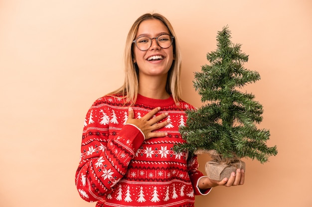 Jovem mulher caucasiana segurando uma pequena árvore de Natal isolada em um fundo bege ri alto, mantendo a mão no peito.
