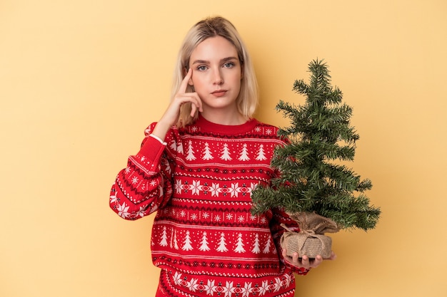 Jovem mulher caucasiana segurando uma pequena árvore de Natal isolada no fundo amarelo, apontando o templo com o dedo, pensando, focada em uma tarefa.