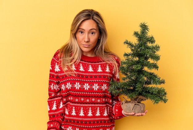 Jovem mulher caucasiana segurando uma pequena árvore de Natal isolada no fundo amarelo encolhe os ombros e abre os olhos confusos.