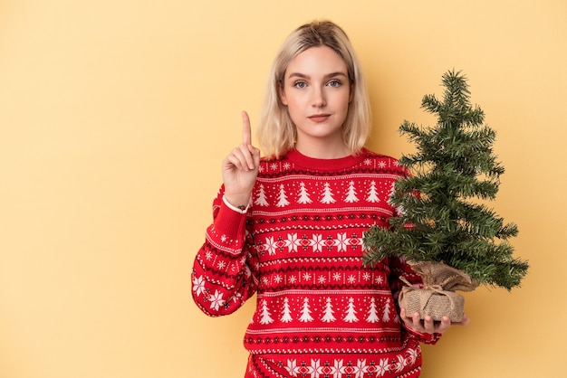 Jovem mulher caucasiana segurando uma pequena árvore de Natal isolada no fundo amarelo, mostrando o número um com o dedo.