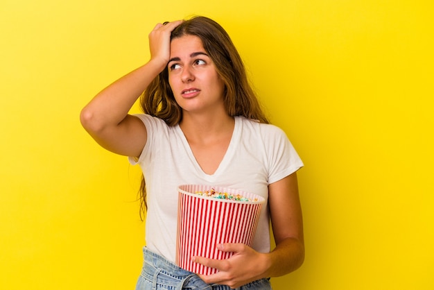 Jovem mulher caucasiana, segurando uma pipoca isolada em um fundo amarelo, sendo chocada, ela se lembrou de uma reunião importante.