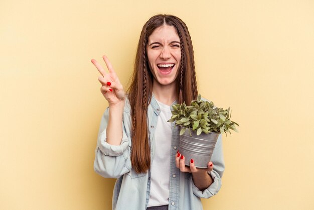 Jovem mulher caucasiana segurando uma planta isolada em fundo amarelo alegre e despreocupada, mostrando um símbolo de paz com os dedos