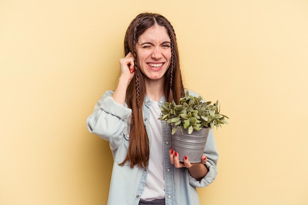 Jovem mulher caucasiana segurando uma planta isolada em fundo amarelo, cobrindo os ouvidos com as mãos