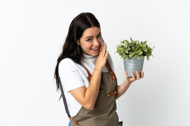 Jovem mulher caucasiana segurando uma planta isolada na parede branca e sussurrando algo