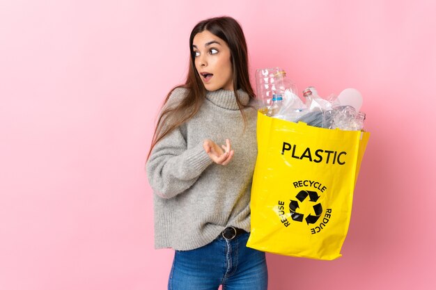 Jovem mulher caucasiana segurando uma sacola cheia de garrafas plásticas para reciclar isolada em um fundo rosa com expressão de surpresa enquanto olha para o lado