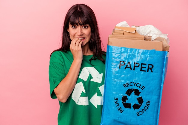 Jovem mulher caucasiana segurando uma sacola de papelão reciclado isolada no fundo rosa, roendo as unhas, nervosa e muito ansiosa.