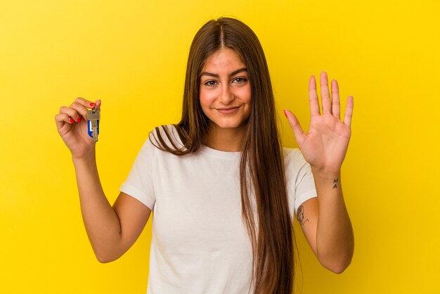 Jovem mulher caucasiana, segurando uma tecla de casa isolada na parede amarela, sorrindo alegre mostrando o número cinco com os dedos.