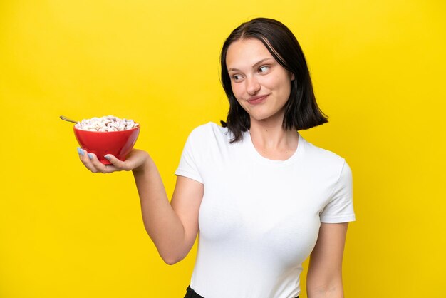 Jovem mulher caucasiana segurando uma tigela de cereais isolada em fundo amarelo com expressão feliz