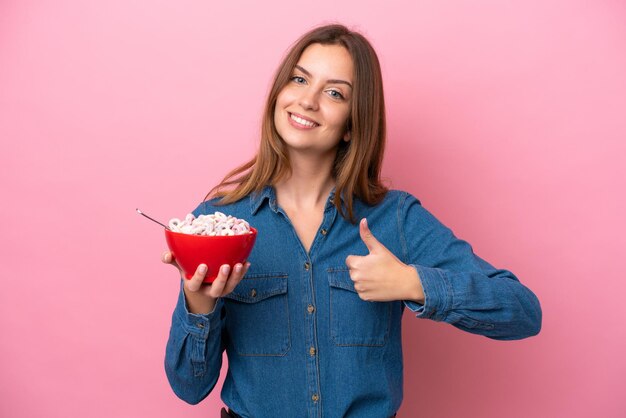 Jovem mulher caucasiana segurando uma tigela de cereais isolada em fundo rosa com polegares para cima porque algo bom aconteceu