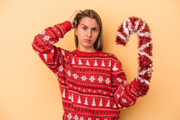 Jovem mulher caucasiana, segurando uma vara de Natal grande isolada em um fundo amarelo, sendo chocada, ela se lembrou de uma reunião importante.