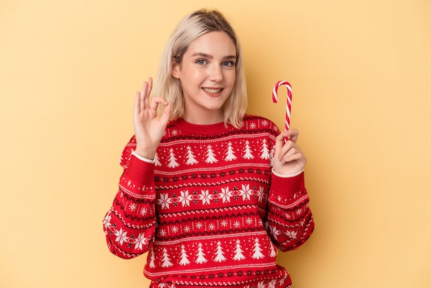 Jovem mulher caucasiana segurando uma vara de Natal isolada em fundo amarelo, alegre e confiante, mostrando um gesto ok.